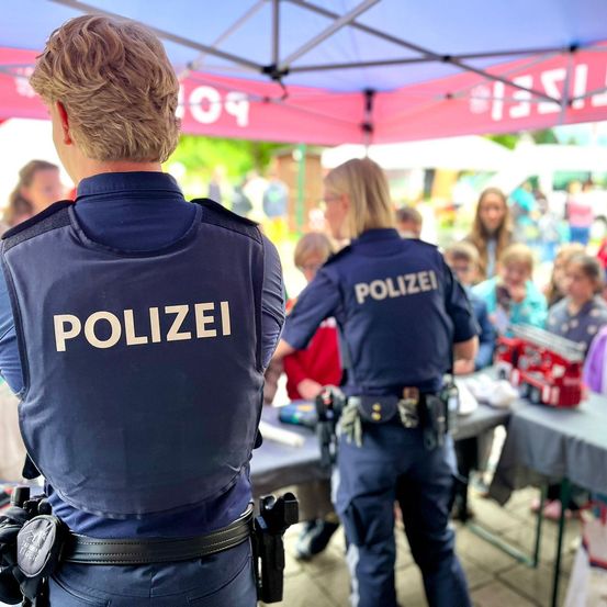 Zwei Polizisten stehen unter einem Zelt bei einer Veranstaltung im Freien, mit einer Menschenmenge im Hintergrund.