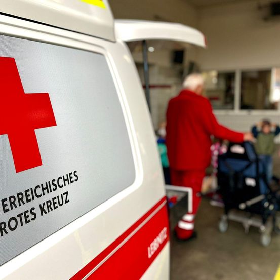 Ein Nahaufnahme der Seite eines Rettungswagens mit einem roten Kreuz und dem Text 'Rotes Kreuz' in Deutsch. Eine Person in Rot schiebt im Hintergrund einen Kinderwagen.