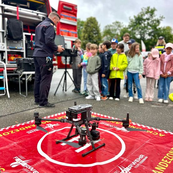 Ein Feuerwehrmann demonstriert ein Drohnenmodell einer Gruppe von Kindern auf einem roten Teppich vor einem Feuerwehrwagen.