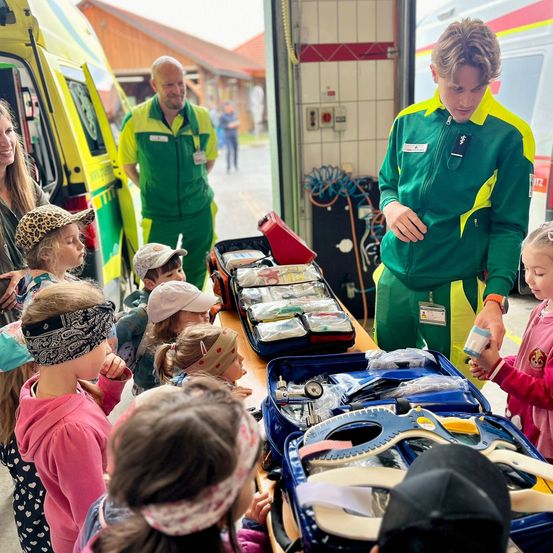 Eine Gruppe von Kindern und Erwachsenen versammelt sich um einen offenen Krankenwagen, wobei ein Sanitäter ihnen medizinische Geräte zeigt.