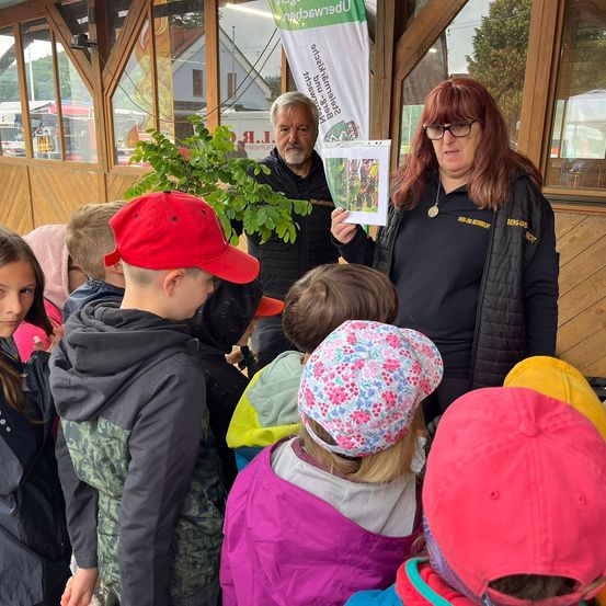 Eine Gruppe von Kindern und zwei Erwachsenen stehen um eine Frau, die eine Pflanze und ein Papier in der Hand hält. Die Frau erklärt den Kindern etwas. Die Kinder tragen Mützen und Jacken. Dahinter steht eine Holzkonstruktion mit einem Banner.