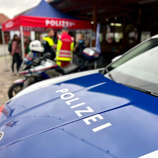 Ein Nahaufnahme eines blauen Polizeiwagens mit dem Wort 'POLIZEI' auf der Motorhaube, geparkt vor einem roten Zelt mit umstehenden Personen.