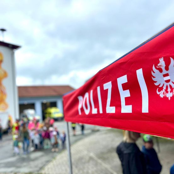 Ein rotes Banner mit dem Wort 'POLIZEI' in weißen Buchstaben wird vor einem Gebäude mit einem Wandbild gezeigt. Im Hintergrund steht eine Menschenmenge, einschließlich Kinder, in der Nähe eines Feuerwehrwagens. Der Himmel ist bewölkt.