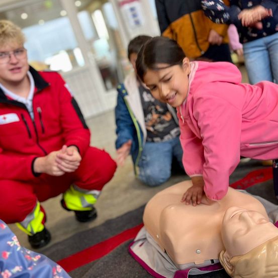Ein junges Mädchen in einer pinken Jacke übt CPR an einem Mannequin, während ein Mann in einem roten Uniform zuschaut.