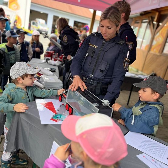 Eine Polizistin zeigt zwei kleinen Jungen ein Spielzeugauto bei einer Veranstaltung. Die Jungen tragen Mützen und Jacken.