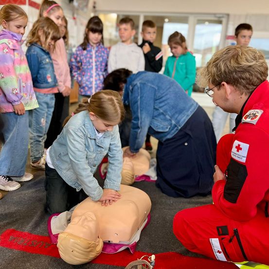 Eine Gruppe von Kindern und Erwachsenen steht um eine CPR-Puppe herum. Ein junges Mädchen übt CPR, während ein Mann in einem roten Uniform zuschaut.