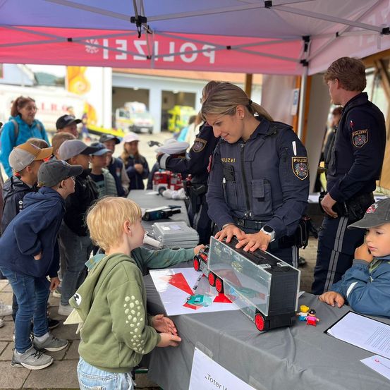 Eine Polizistin zeigt einem kleinen Jungen einen Spielzeuglastwagen bei einer Veranstaltung im Freien unter einem Zelt. Andere Kinder und Erwachsene schauen zu.