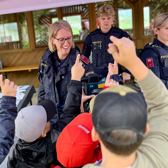 Eine lächelnde Polizistin umgeben von Kindern und zwei männlichen Beamten, einer hält eine Kamera, in einer sonnigen Außenumgebung.