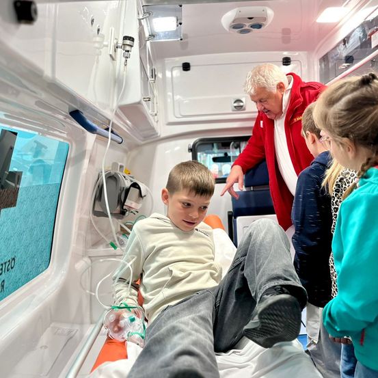 Ein Mann in einem roten Jackett erklärt etwas einem Jungen, der auf einer Trage in einem Krankenwagen liegt. Der Junge hat eine Sauerstoffmaske und schaut den Mann an.