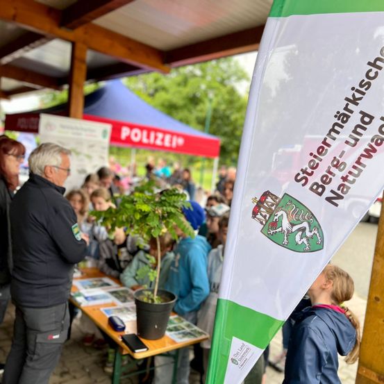 Eine Gruppe von Menschen, einschließlich Kindern, versammelt sich um einen Tisch mit einem Topfpflanzen und Informationsmaterialien. Ein Flagge mit einem Wappen und Text ist im Vordergrund sichtbar. Hinter dem Tisch steht ein Polizeizelt mit einer Menschenmenge.