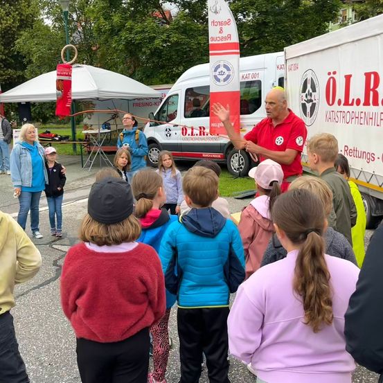 Ein Mann in einem roten Shirt hält eine Präsentation für eine Gruppe von Kindern vor einem weißen Krankenwagen mit dem Text O.L.R. auf der Seite.