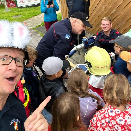 Eine Gruppe von Feuerwehrleuten interagiert mit Kindern, einer trägt einen Hasenohrenhut und der andere einen gelben Helm. Sie sind um einen Feuerlöscher versammelt.