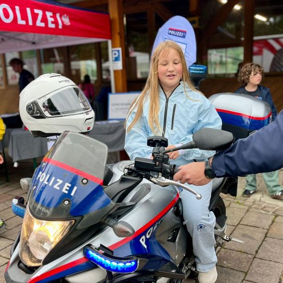 Ein junges Mädchen sitzt auf einem Polizeimotorrad, mit einem Helm auf dem Lenker. Ein Polizist steht neben ihr und hält den Lenker. Die Szene spielt sich im Freien ab, mit einem Polizeizelt im Hintergrund.