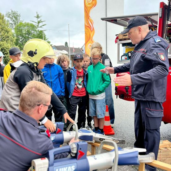 Ein Feuerwehrmann demonstriert ein Werkzeug für eine Gruppe von Kindern, mit einem Feuerwehrwagen und Sicherheitskegeln im Hintergrund.
