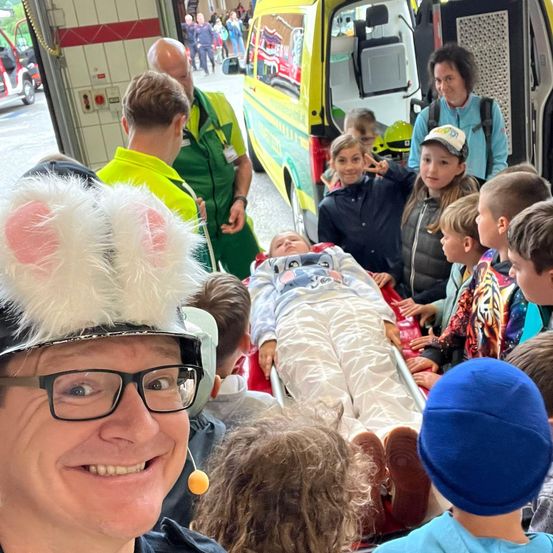 Eine Gruppe von Kindern und Erwachsenen versammelt sich um einen Krankenwagen. Eine Person in einer Hasenmütze lächelt in die Kamera. Eine Person liegt auf einer Trage.