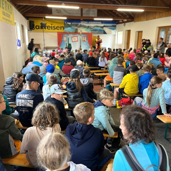 Eine Gruppe von Menschen, wahrscheinlich Schüler, sitzt in einem Raum, möglicherweise einem Klassenzimmer, und schaut sich eine Aufführung auf einer Bühne an. Die Bühne hat ein Banner mit Text und einer Person davor. Der Raum hat Holzbänke, eine Holzdecke und eine Wand mit einem Fenster.