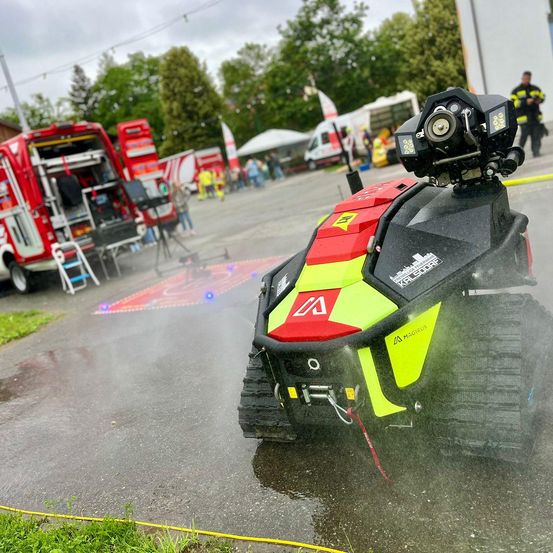 Ein rotes und gelbes Roboterfahrzeug mit einer Kamera oben sprüht Wasser auf die Straße. Im Hintergrund sind Feuerwehrwagen und Menschen zu sehen.