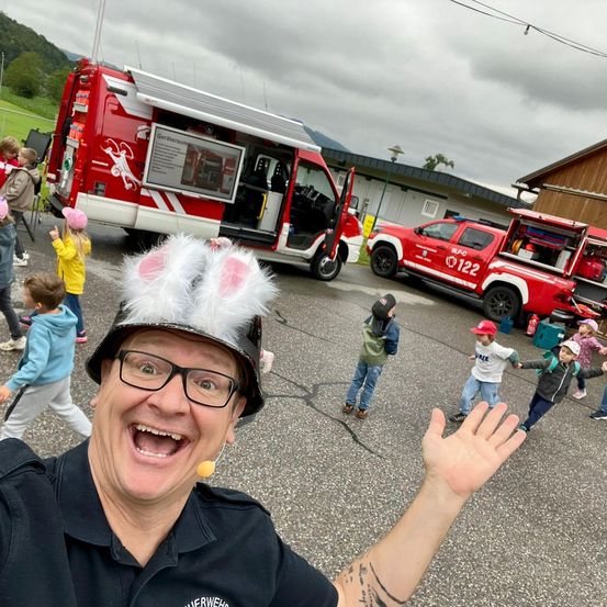 Ein Mann in einer Feuerwehruniform und einem Hasenohrenhut winkt zur Kamera, während eine Gruppe von Kindern und Feuerwehrwagen im Hintergrund zu sehen sind.