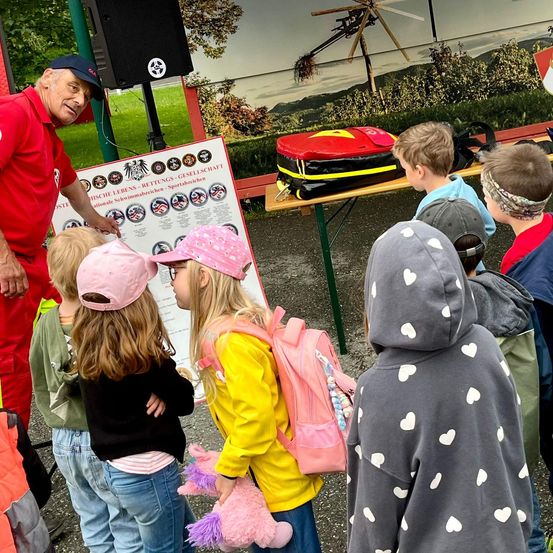 Eine Gruppe von Kindern, einige mit Mützen, stehen um einen Mann in einem roten Uniform herum. Sie betrachten ein Plakat mit Abzeichen und Text. In der Nähe steht ein Tisch mit einem roten und schwarzen Rucksack.