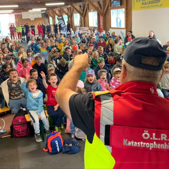 Eine große Gruppe von Kindern und Erwachsenen ist in einem Saal versammelt, einige sitzen auf Bänken und andere stehen, mit einem Mann in einer roten Sicherheitsjacke, der eine Sitzung leitet.