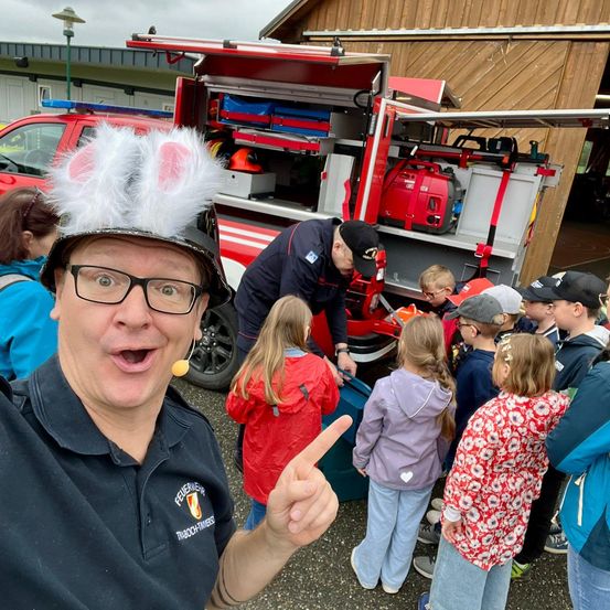 Ein Mann mit einem Feuerwehrmannhut mit Hasenohren macht ein Selfie mit einer Gruppe von Kindern vor einem Feuerwehrwagen.