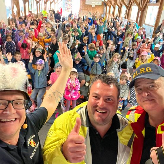 Eine Gruppe von Menschen, einschließlich Kindern und Erwachsenen, versammelt sich in einem Raum und lächelt und winkt. Drei Erwachsene stehen vorne, einer davon gibt einen Daumen hoch. Die Kinder tragen Hüte und Jacken, und einige heben ihre Hände. Der Raum hat Holzbalken und Glasfenster.