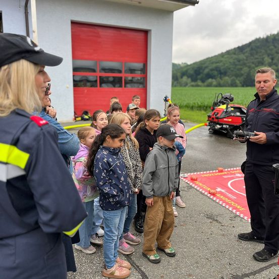 Eine Gruppe von Kindern und Erwachsenen ist um einen Feuerwehrmann in Uniform versammelt, der ihnen zuhört, während er eine Fernbedienung in der Hand hält. Hinter ihnen befindet sich eine Feuerwache mit einer roten Tür und einem Feuerwehrauto.
