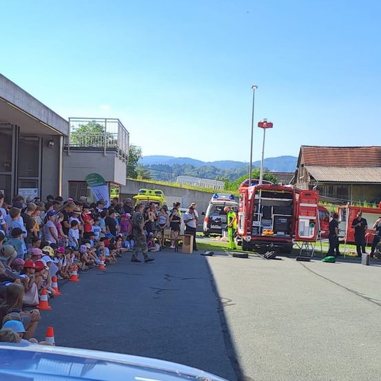 Eine Menschenmenge, darunter Kinder, versammelt sich um einen Feuerwehrwagen und einen Krankenwagen. Die Fahrzeuge sind am Straßenrand geparkt, mit einem Gebäude und Bergen im Hintergrund.