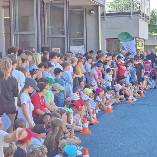 Eine große Menschenmenge, darunter auch Kinder, ist im Freien versammelt, wahrscheinlich für eine Veranstaltung. Viele tragen Hüte und einige halten Papiere. Verkehrskegel säumen das Gebiet.