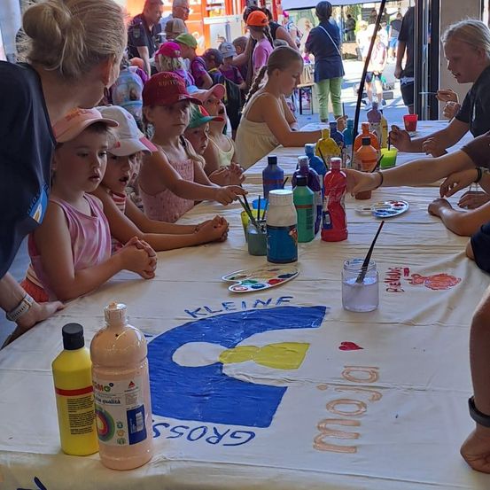 Eine Gruppe von Kindern malt auf einem großen Tisch mit Flaschen und Pinseln. Sie sitzen um den Tisch herum, der mit einem weißen Tuch und einem Logo darauf bedeckt ist.