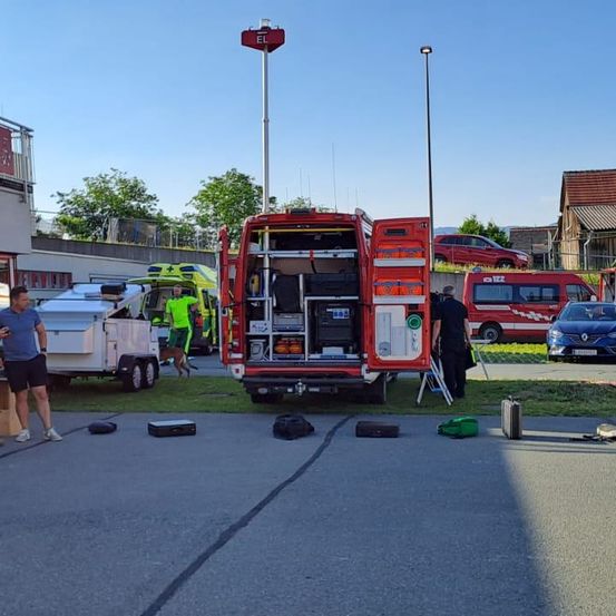 Ein roter Lieferwagen mit offenen hinteren Türen ist am Straßenrand geparkt. Ein Mann steht in der Nähe, und eine Person geht auf den Lieferwagen zu. Ein weißer Anhänger ist hinter dem Lieferwagen geparkt, und eine Person steht in der Nähe. Ein Auto ist auf der rechten Seite geparkt.