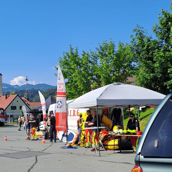 Eine Gruppe von Personen in Sicherheitsausrüstung versammelt sich um ein Zelt mit einem Tisch und verschiedenen Ausrüstungsgegenständen, möglicherweise für eine Schulungsveranstaltung. Im Hintergrund befinden sich Gebäude, Bäume und Berge.