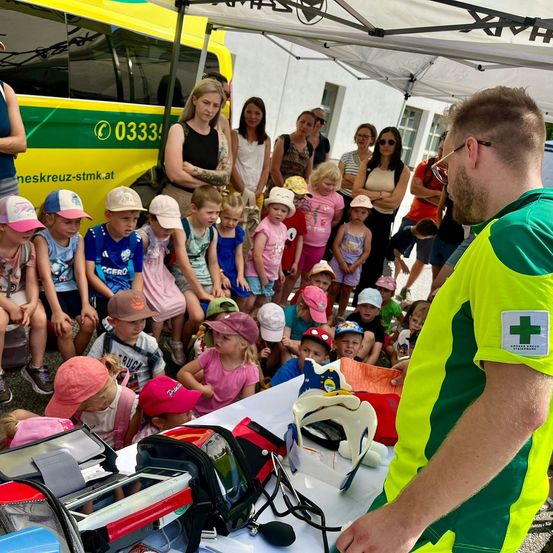 Eine Gruppe von Kindern und Erwachsenen versammelt sich unter einem Zelt, ein Mann in einem grün-gelben Uniform steht an einem Tisch mit medizinischen Geräten.