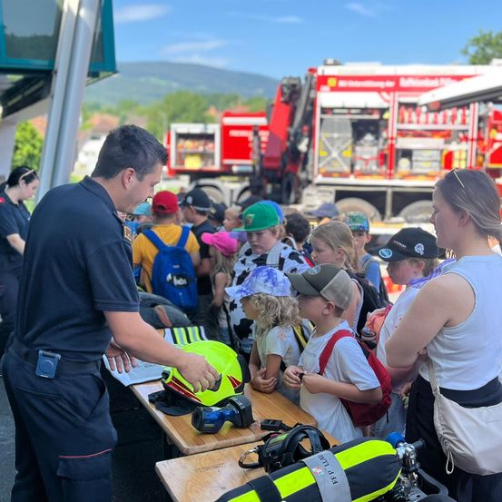 Eine Gruppe von Kindern und Erwachsenen versammelt sich um einen Tisch mit Helmen und anderer Feuerwehrausrüstung, mit einem Feuerwehrwagen im Hintergrund.