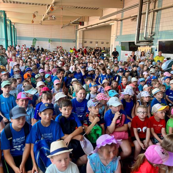 Eine große Gruppe von Kindern in blauen und weißen Uniformen, Hüten und Brillen sitzt in einem überfüllten Innenraum, möglicherweise einer Sporthalle. Einige tragen Rucksäcke.