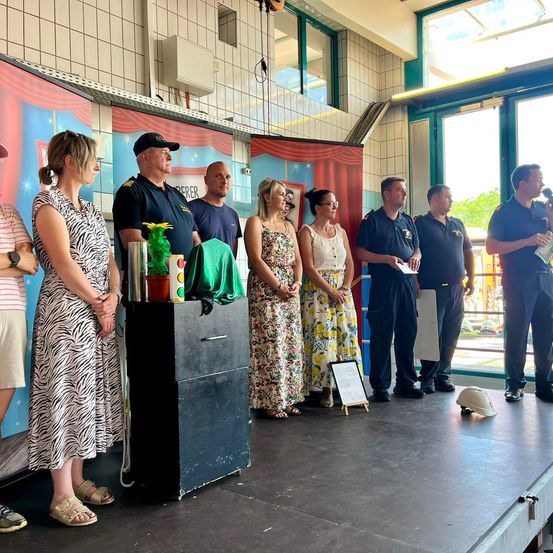 Eine Gruppe von Menschen steht auf einer Bühne mit einem Hintergrund. Ein Mann in blauer Uniform hält ein Mikrofon und scheint zu sprechen. Eine Frau in einem Zebra-Druckkleid steht neben einer schwarzen Box mit einem grünen Tuch obenauf.