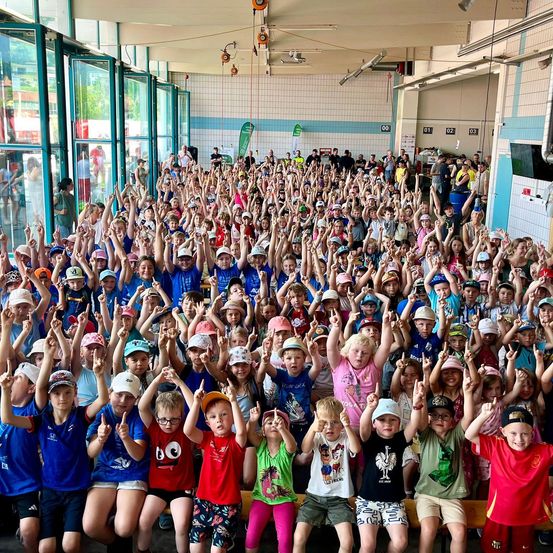 Eine große Gruppe von Kindern in einer Turnhalle, alle mit Mützen und erhobenen Händen, einige mit Friedenszeichen. Die Kinder sind in verschiedenen Farben gekleidet und scheinen an einer Veranstaltung teilzunehmen.
