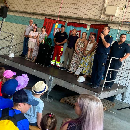 Eine Gruppe von Erwachsenen und Kindern steht auf einer Bühne, wahrscheinlich für eine Veranstaltung. Die Erwachsenen stehen, während die Kinder auf dem Boden sitzen. Die Bühne hat einen Holzboden und ein Metallgeländer. Die Erwachsenen tragen Uniformen, Hüte und Schuhe. Die Kinder tragen Hüte und Schuhe. Die Bühne hat einen Lautsprecher, eine schwarze Box und eine Tafel.