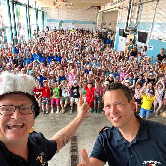 Zwei Männer in Feuerwehruniformen posieren für ein Foto mit einer großen Menge von Kindern in einer Turnhalle. Die Kinder tragen Mützen und einige heben ihre Hände.