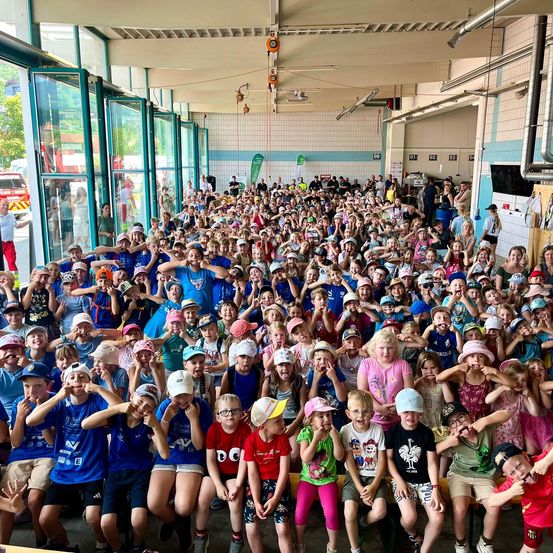 Eine große Gruppe von Kindern und Erwachsenen versammelt sich in einem großen Innenraum, wahrscheinlich einer Sporthalle, mit Hüten und einigen, die Handgesten machen. Die Kinder sitzen auf dem Boden, während andere stehen. Der Raum hat Glastüren und Fenster, und es stehen Menschen draußen.