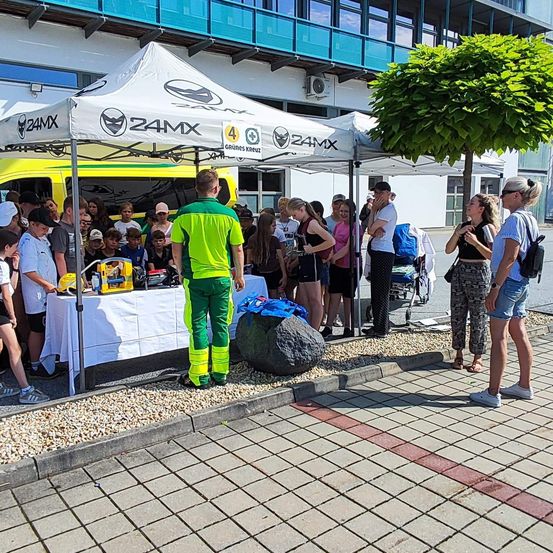 Eine Gruppe von Menschen versammelt sich um ein Zelt mit einem gelben Van dahinter. Ein Mann in grüner und gelber Kleidung steht vor einem Tisch mit Gegenständen darauf.
