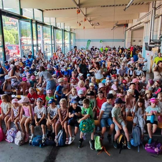 Eine große Gruppe von Kindern und Erwachsenen versammelt sich in einem Saal, einige sitzen auf Bänken und andere stehen. Viele tragen Hüte und haben Rucksäcke dabei. Der Saal hat Glaswände und Türen, und draußen sind Feuerwehrwagen zu sehen.
