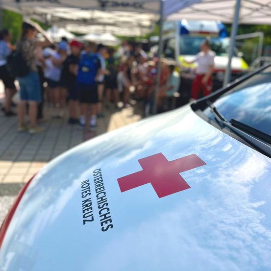 Ein weißer Krankenwagen mit einem roten Kreuz auf der Tür steht vor einer Menschenmenge unter einem Zelt.
