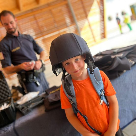 Ein junger Junge mit Helm und Rucksack steht neben einem Mann in Uniform, möglicherweise bei einer Veranstaltung.