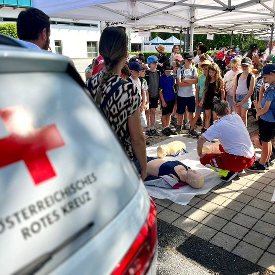 Eine Gruppe von Menschen, einschließlich Kindern, ist um einen Krankenwagen des Roten Kreuzes versammelt. Eine Erste-Hilfe-Demonstration findet mit einer Puppe auf dem Boden statt.