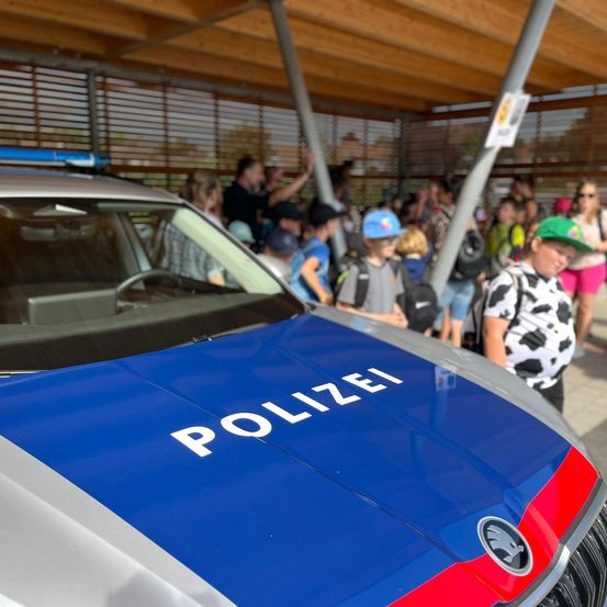 Ein Polizeiauto mit dem Wort 'Polizei' auf der Motorhaube, dahinter steht eine Menschenmenge, einige tragen Hüte und haben Rucksäcke dabei.