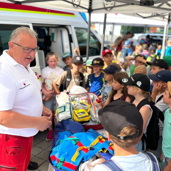 Ein Mann in einem weißen Hemd und roten Hosen steht vor einem Tisch mit medizinischen Geräten und spricht eine Gruppe von Kindern in Mützen an.