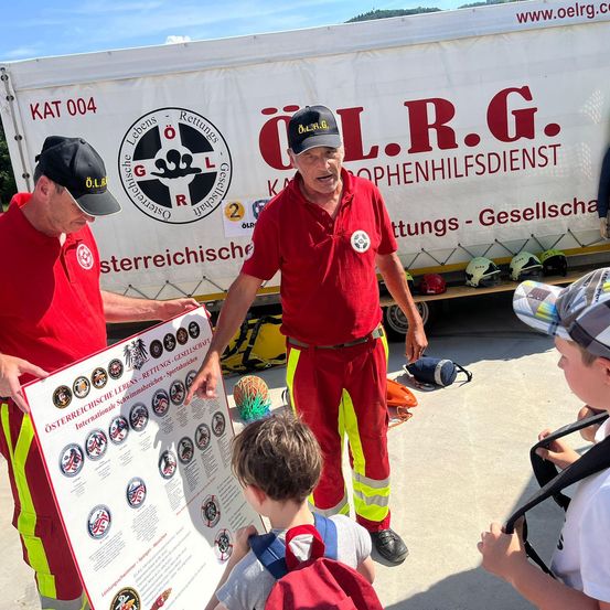 Zwei Männer in roten Uniformen zeigen einem Kind und einer anderen Person ein Poster mit verschiedenen Abzeichen. Der Poster hat den Text 'Osterreichische Lebens-Rettungs-Gesellschaf'. Dahinter steht ein weißer LKW mit dem Text 'O.L.R.G.' und 'Kat 004'. Auf dem LKW befinden sich Helme und andere Ausrüstung.