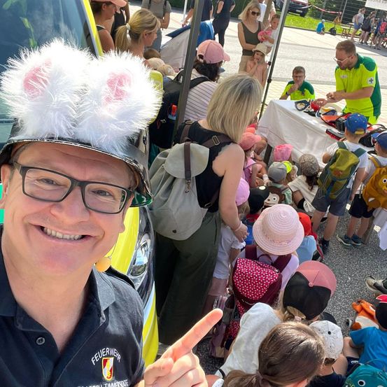 Ein Feuerwehrmann in einem Hasenohrenhut posiert für ein Foto mit einer Gruppe von Kindern und Erwachsenen bei einer Veranstaltung im Freien.