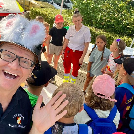 Eine Gruppe von Kindern und Erwachsenen versammelt sich draußen. Ein Feuerwehrmann in einem Helm und mit Brille lächelt. Eine Frau in einem weißen Shirt und roten Hosen steht vor den Kindern.